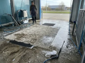 Mokry dywan leży na posadzce myjni samochodowej obok piany i lancy ciśnieniowej, pokazując ryzyko takiego prania.